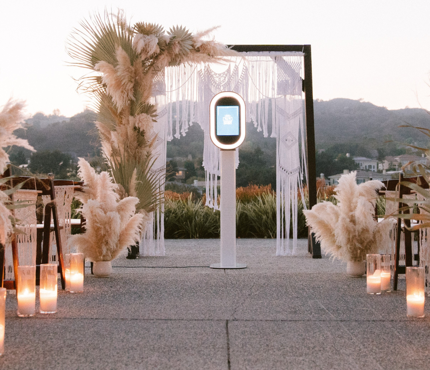 Outdoor setting with palms, dried feathery leaves, chairs, lamps and a decorative gazebo with tassels and a photo booth in the center. Along with a mountainous background.