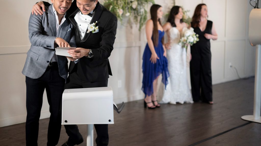 Two men looking at photo prints and laughing. Three women in the background posing in front of a photo booth.