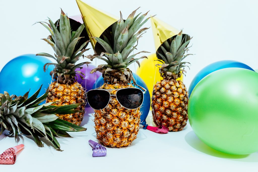 Pineapples, one wearing sunglasses, colorful balloons, party hats and party blowers.